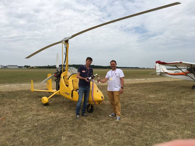 Deux hommes debout à côté d'un ULM jaune sur un terrain d'aviation, l'un tenant un trophée.