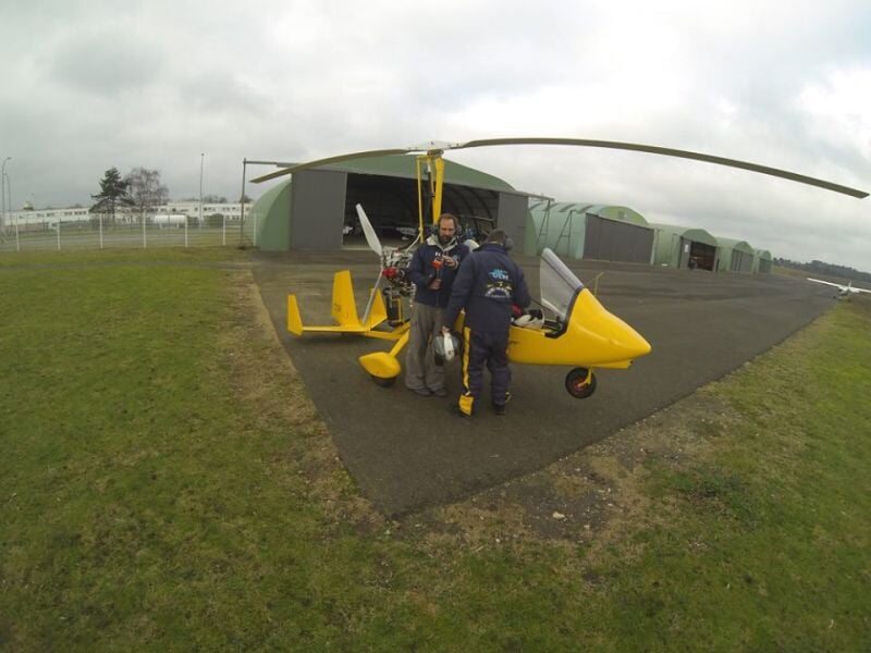 Trois personnes près d'un ULM jaune sur une piste d'aviation, avec des hangars en arrière-plan.