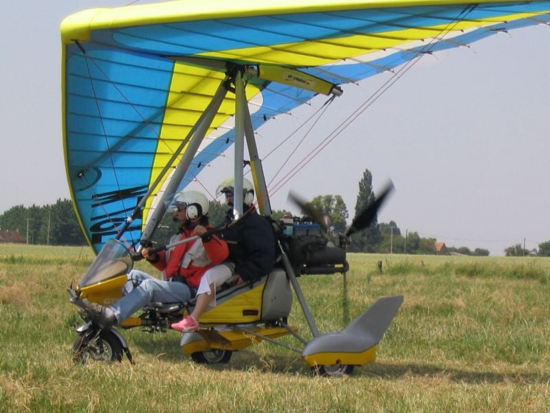 ULM jaune au sol sur un champ, avec deux personnes équipées de casques prêtes à décoller.