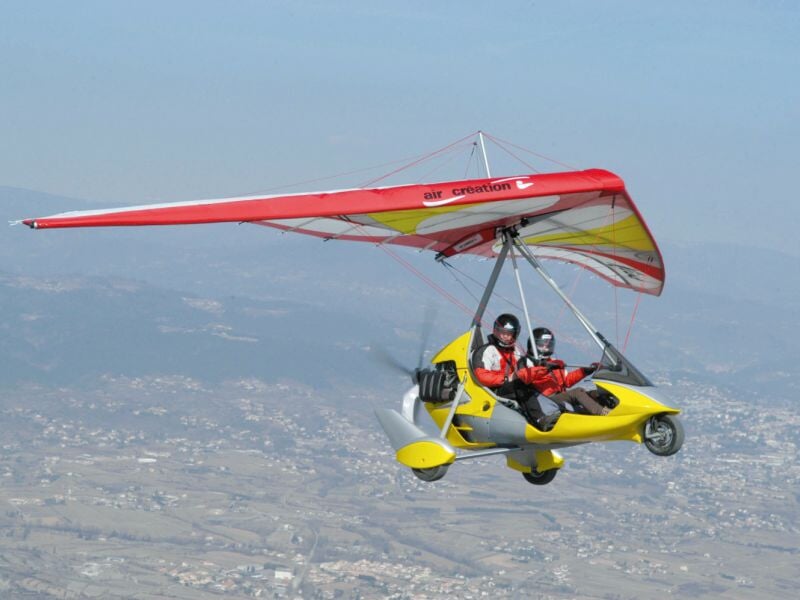 ULM rouge et jaune en vol avec deux passagers portant casque, survolant un paysage montagneux.