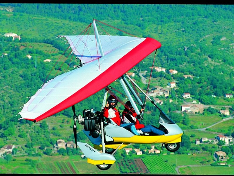 ULM rouge et blanc en vol avec deux personnes à bord, survolant une zone verte et habitée.