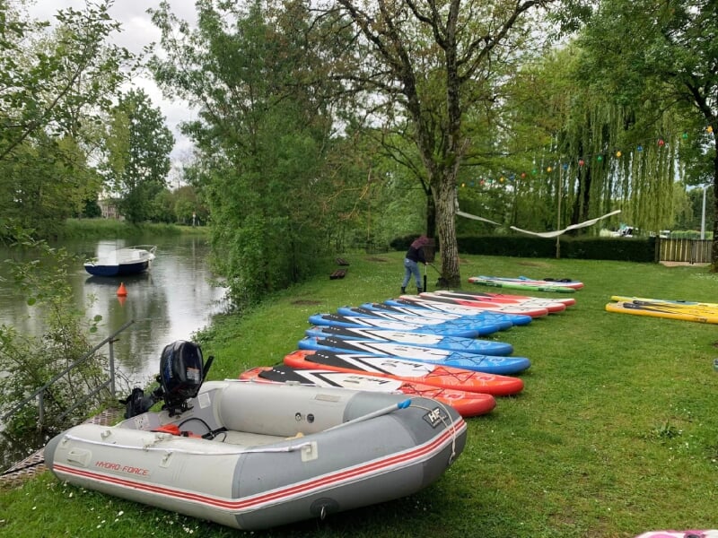 Bateaux gonflables et paddles multicolores rangés près d'une rivière calme, des décorations festives suspendues dans les arbres environnants.