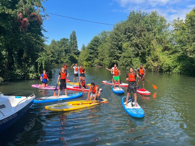 Groupe de personnes pratiquant le paddle sur une rivière bordée de végétation luxuriante, tous portant des gilets de sauvetage orange et pagayant sous un ciel bleu.