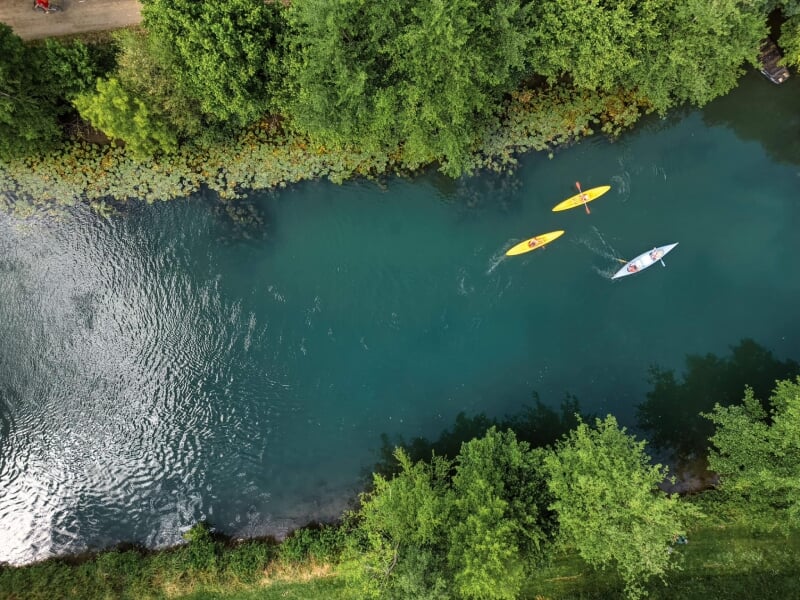 Vue aérienne de trois kayakistes naviguant sur une rivière sinueuse, entourée de végétation luxuriante et de nénuphars flottants.