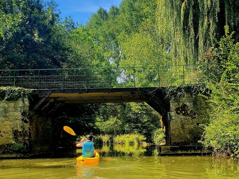 Kayakiste pagayant sous un pont de pierre, entouré de végétation dense et de grands arbres, par une journée ensoleillée.