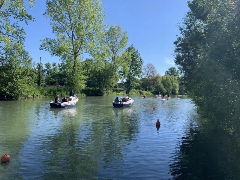 Plusieurs bateaux naviguant sur une rivière par une journée ensoleillée, bordée d'arbres et de bouées oranges. Ambiance détendue et estivale.
