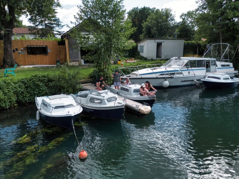 Bateaux amarrés le long d'un quai, entourés de végétation et de bâtiments en arrière-plan. Des personnes discutent sur les ponts des bateaux, ambiance conviviale et estivale.
