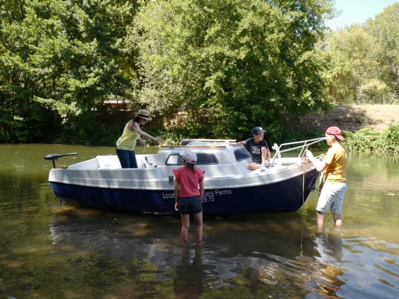 Location de bateaux sans permis en rivière, une famille se préparant à embarquer. Enfants et adultes en tenue estivale, cadre naturel verdoyant et ensoleillé.