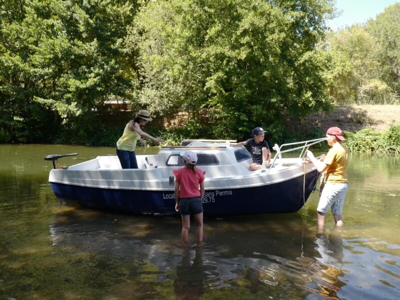 Location de bateaux sans permis en rivière, une famille se préparant à embarquer. Enfants et adultes en tenue estivale, cadre naturel verdoyant et ensoleillé.