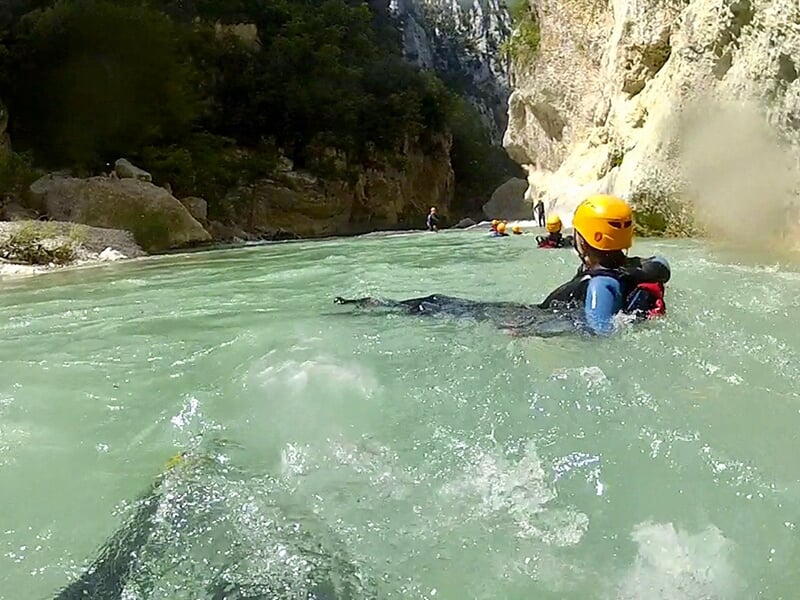 Un sportif vêtu d'une combinaison néoprène noire et d'un casque jaune se laisse porter par le courant d'un torrent de montagne.