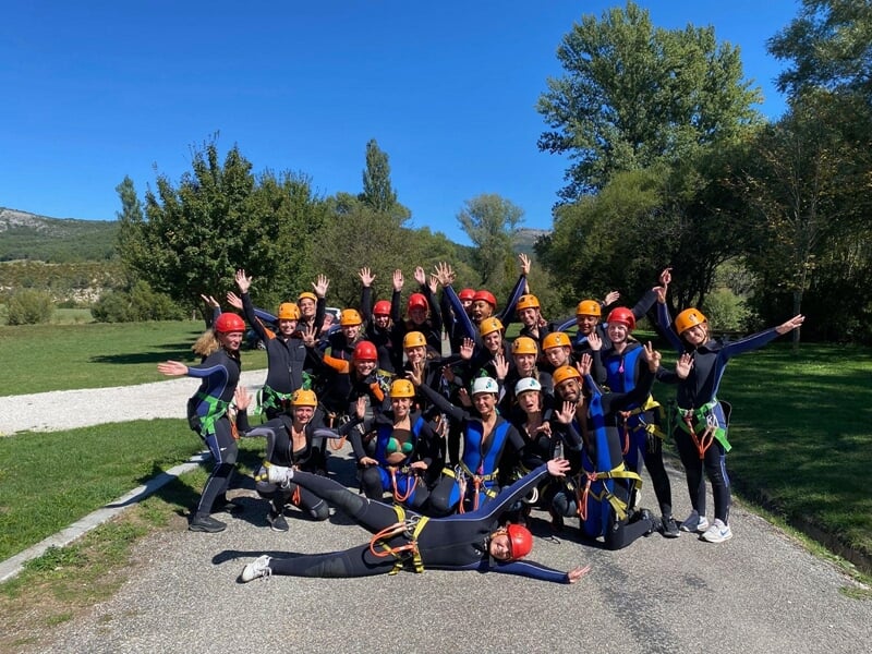 Un groupe de sportifs casqués et en combinaison néoprène posent en levant les bras avec des arbres et un ciel bleu en arriètre-plan