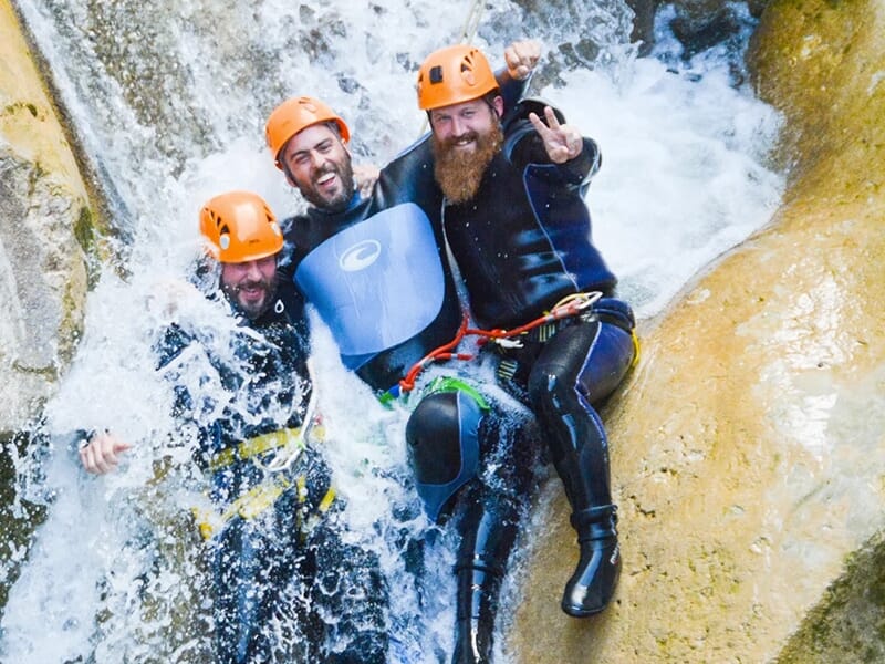 Trois amis se tenant par les épaules descendent un toboggan en riant lors d'une sortie de canyoning