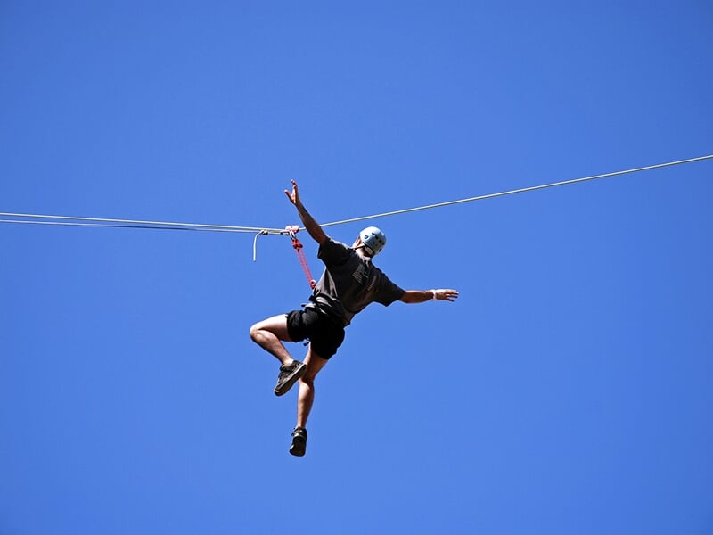 Un homme en short et les bras tendus en plein milieu d'une descente en tyrolienne avec en toile de fond le ciel bleu