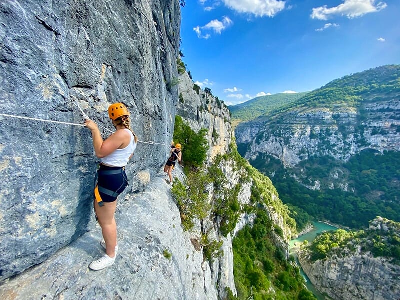 Deux jeunes femmes portant un casque progressent à flanc de falaise suspendues à une ligne de vie