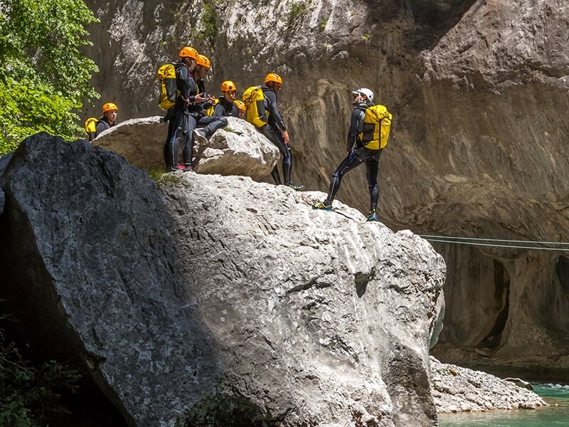 Un groupe de canyoneurs portant des casques, des combinaisons néoprènes et des sacs étanches sont postés sur un rocher dans un canyon.