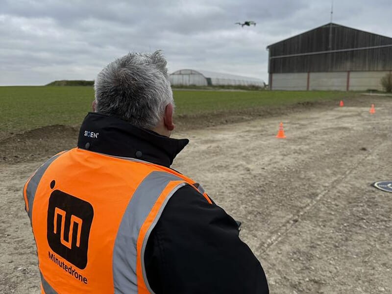 Un homme portant un chasuble de sécurité orange fait voler un drone au dessus d'une piste d'atterrissage installée près d'un hangar