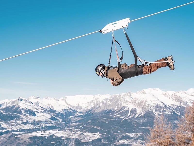 En homme portant un casque en plein au milieu d'une descente en tyrolienne avec les Alpes enneigées en toile de fond