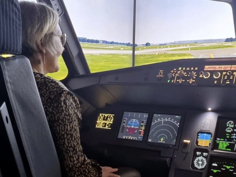 Femme aux cheveux clairs et lunettes assise dans un simulateur de cockpit, regardant un paysage d’aéroport projeté devant elle.