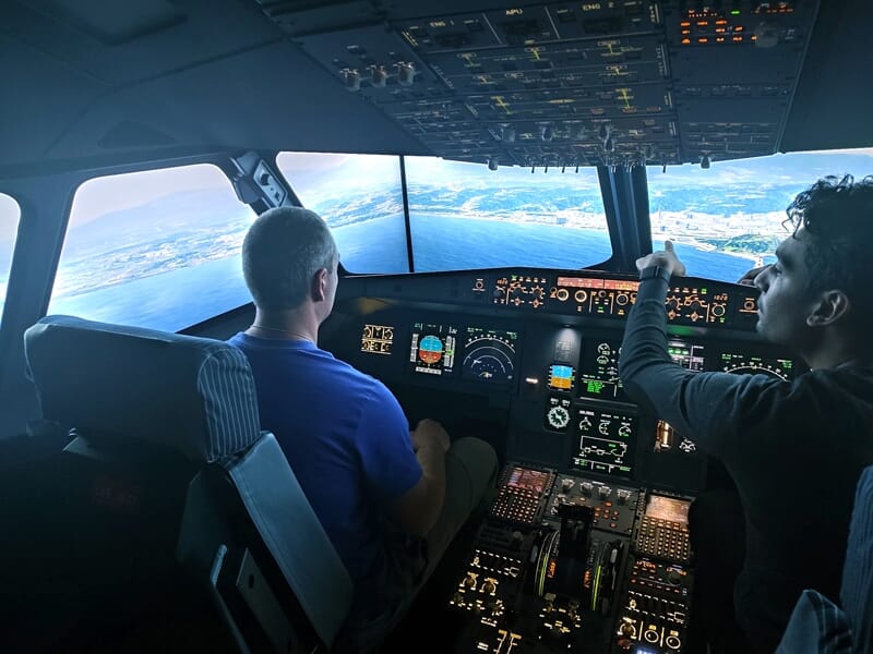 Deux hommes dans un simulateur de cockpit en vol, l’un en t-shirt bleu aux commandes, l’autre en pull gris pointant un écran.