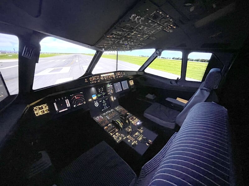 Poste de pilotage d’un simulateur de vol avec sièges capitonnés, tableau de bord illuminé et vue sur une piste d’atterrissage par les fenêtres avant.