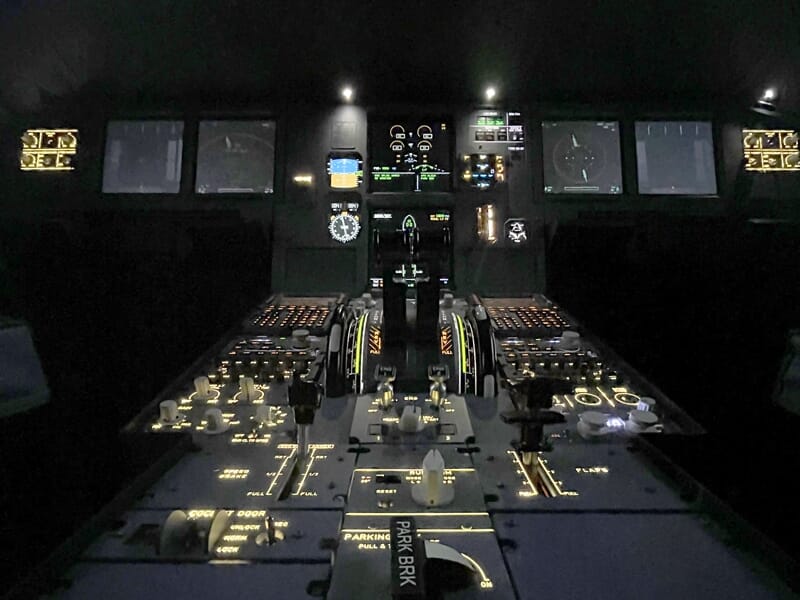 Vue centrée sur les manettes de gaz et les panneaux de commande inférieurs du cockpit, avec nombreux voyants et interrupteurs allumés.
