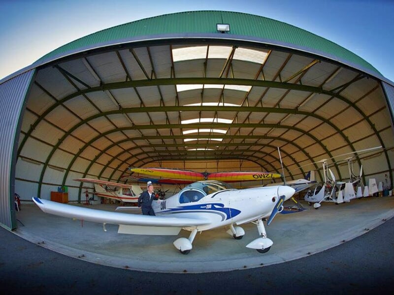 Un pilote d'avion en tenue verte se tient devant un ULM Pioneer 200 à l'entrée du hangar d'un aérodrome 