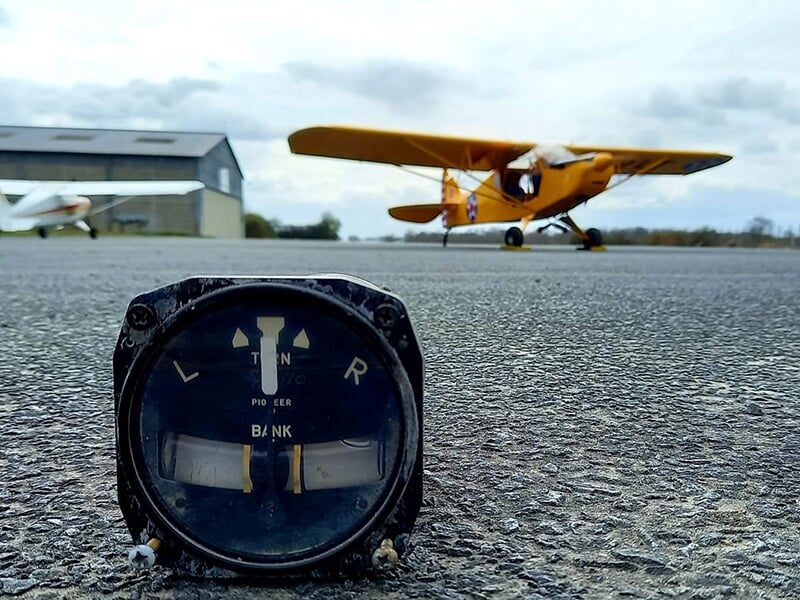 Un altimètre vintage est posé sur la piste d'atterrissage d'un aérodrome avec deux avions et un hangar en arrière-plan