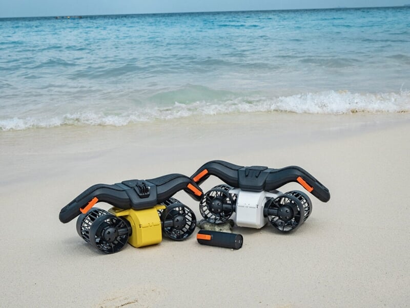 Deux scooters sous-marins posés sur une plage de sable fin avec la mer en toile de fond.