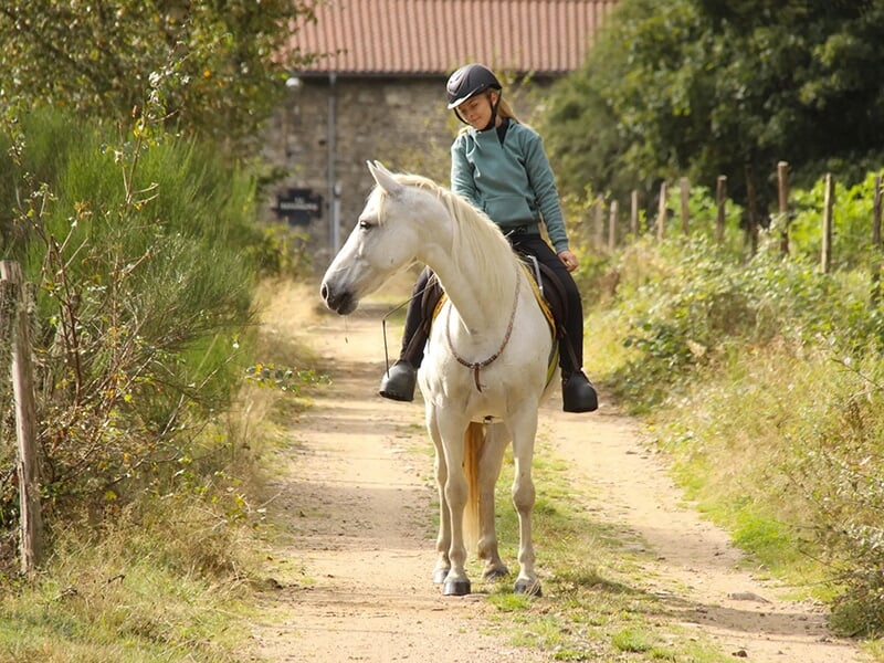 Une jeune fille chevauche un cheval blanc sur un chemin de terre avec une bâtisse en arrière-plan