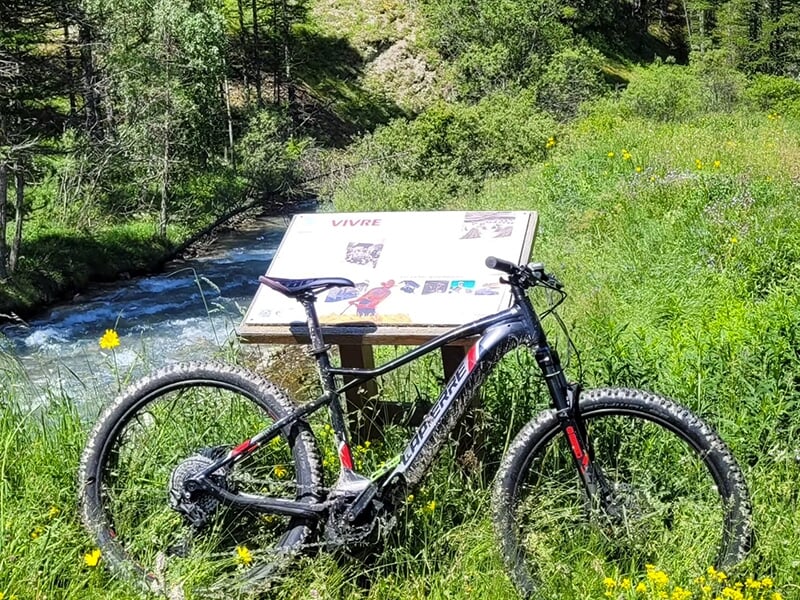 Un vélo électrique tout terrain appuyé sur un panneau touristique en pleine nature devant un torrent de montagne 