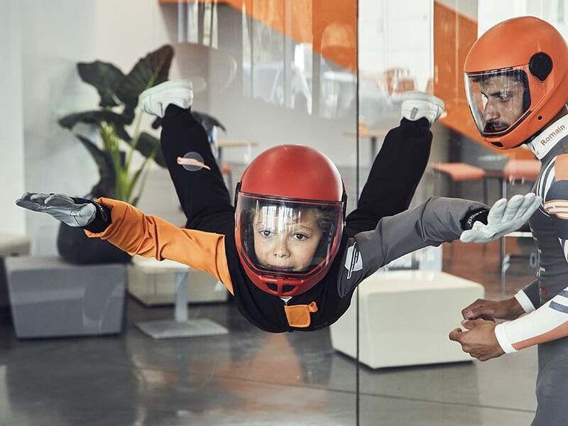 Un jeune enfant équipé d'un casque rouge et d'une combinaison noire et orange, flottant dans une soufflerie intérieure. Un instructeur en combinaison grise l'accompagne pour guider l'enfant dans l'exp