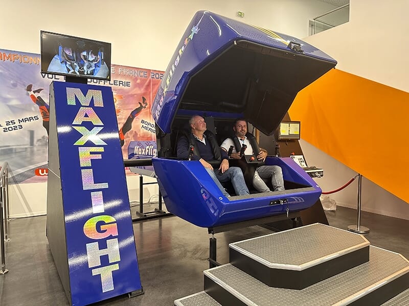 Deux hommes sont assis dans un simulateur de vol bleu, nommé MaxFlight. Ils sont à l'intérieur du simulateur en forme de capsule, prêt à vivre une expérience immersive.