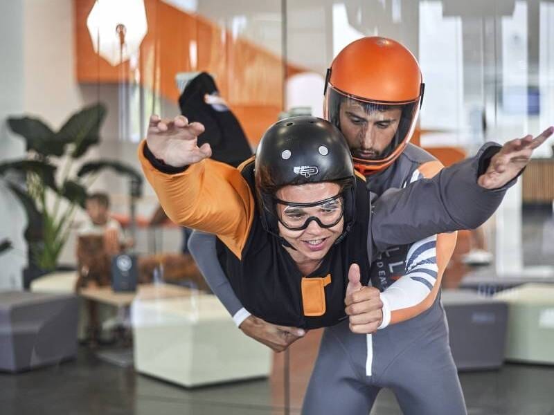 Simulation de chute libre en soufflerie intérieure avec un instructeur. Une personne portant une combinaison noire et orange, un casque et des lunettes de protection est guidée par un moniteur en tenu