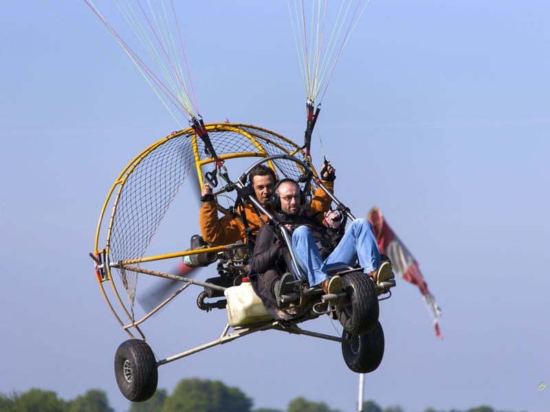 Deux personnes en vol sur un paramoteur, avec l'un des pilotes saluant, sur fond de ciel clair
