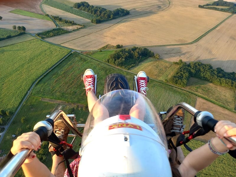 Vue en première personne des pieds d'une personne sur un paramoteur, survolant un paysage de champs verts et de routes