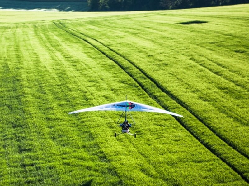 ULM glissant juste au-dessus d'une étendue d'herbe verte, avec des lignes parallèles marquant le terrain, éclairé par une lumière vive, soulignant la précision et la maîtrise du pilotage en milieu rur