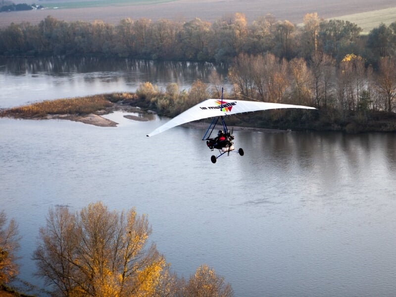 Deltaplane motorisé survolant une rivière paisible au crépuscule, les reflets de l'eau miroitant sous un ciel légèrement brumeux, encadré par une végétation automnale aux tons chauds de jaunes et de v