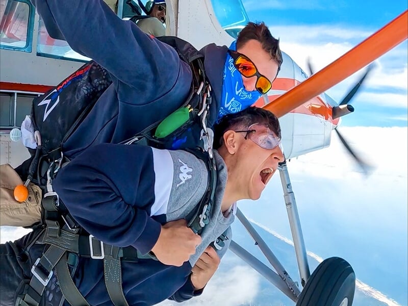 Deux parachutistes en tandem sautant d'un avion en plein ciel, l'un d'eux exprimant une grande excitation.