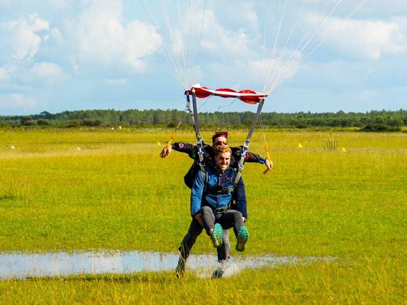 Parachutistes en tandem atterrissant dans un champ verdoyant après un saut, avec le parachute ouvert au-dessus d'eux.