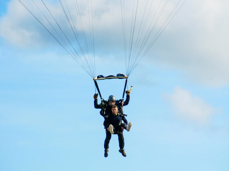 Deux parachutistes en tandem descendant lentement avec le parachute ouvert, souriants, sous un ciel partiellement nuageux.