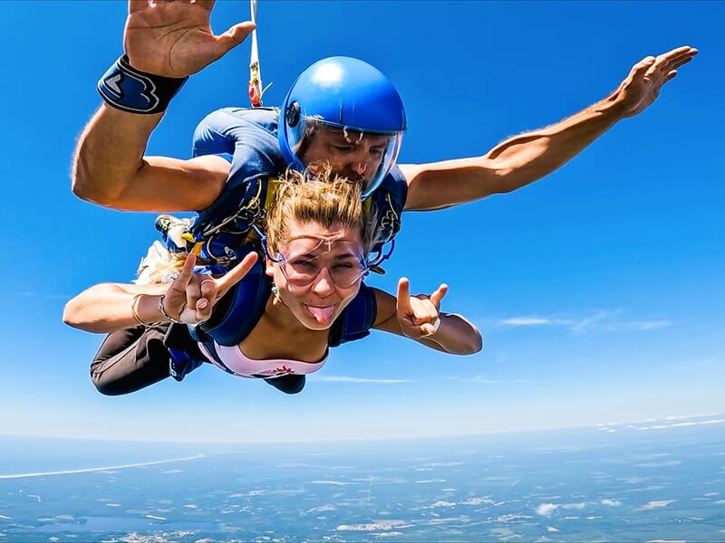 Parachutiste en tandem avec une femme faisant un signe de rock, la langue sortie, en chute libre au-dessus d'un paysage vaste et bleu.