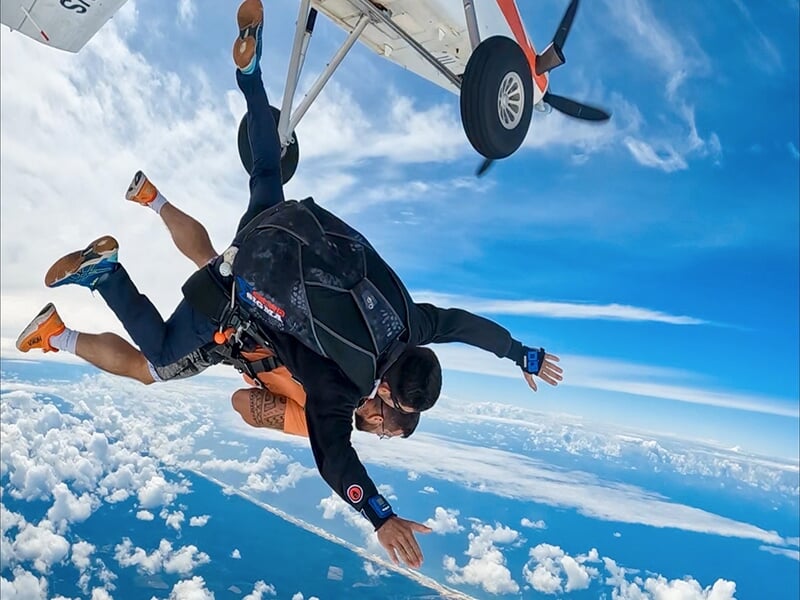 Parachutistes en tandem en pleine chute libre, avec le passager souriant et levant les mains en signe de joie sous un ciel bleu clair.