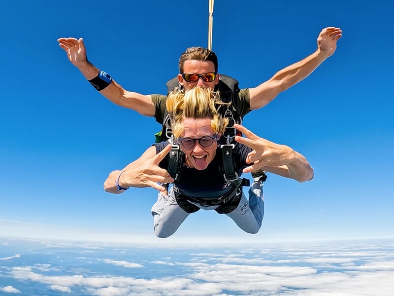 Parachutistes en tandem descendant avec enthousiasme, le passager tirant la langue et faisant des gestes de paix, sous un ciel bleu.