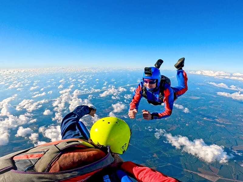 Deux parachutistes en pleine chute libre, l'un en combinaison bleue et rouge, échangeant un poing avec un autre parachutiste portant un casque jaune, au-dessus d'un ciel clair avec des nuages.