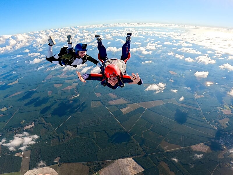 Deux parachutistes chutant côte à côte, l'un portant un casque rouge, sous un ciel dégagé avec un vaste paysage de champs en arrière-plan.
