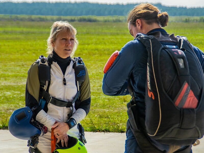 Deux parachutistes en discussion au sol, l'un en combinaison blanche tenant un casque jaune et l'autre ajustant son équipement, dans un environnement de prairie.