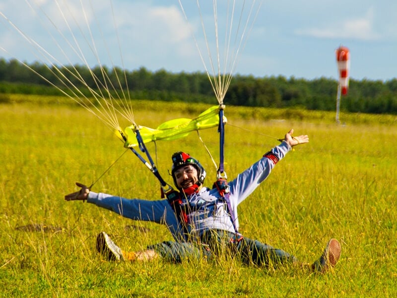 Parachutiste assis dans l'herbe après l'atterrissage, souriant et levant les bras avec le parachute déployé derrière lui.