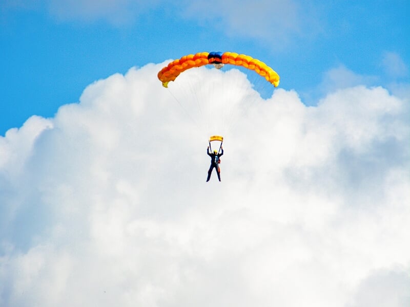Parachutiste descendant sous un parachute jaune et orange vif, flottant doucement avec des nuages en arrière-plan.