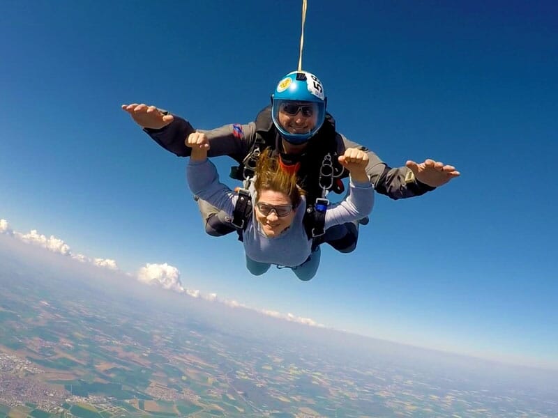 Deux personnes en chute libre, l'instructeur en combinaison noire et bleue tenant la parachutiste par les bras. La parachutiste porte une combinaison grise et des lunettes de protection. Le ciel est d