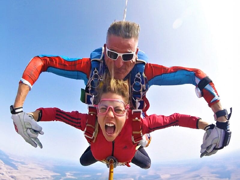 Parachutiste en tandem en chute libre avec un grand sourire et les bras écartés. L'instructeur derrière elle porte des lunettes de soleil blanches et une combinaison bleue et rouge. Le ciel dégagé et 